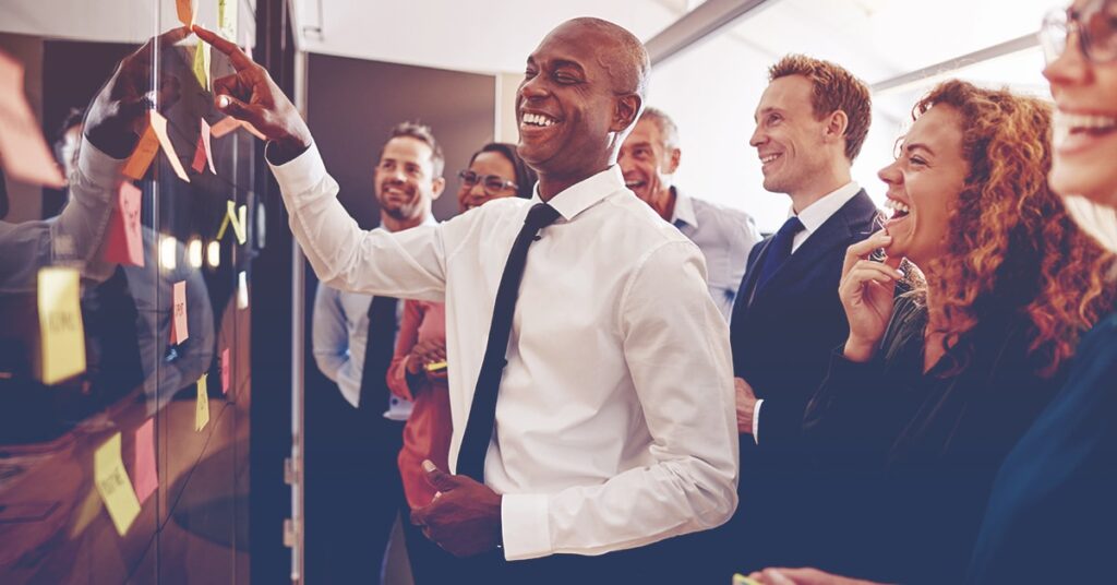 Leader reviewing suggestions from a training session, pointing at a sticky note and laughing as his team laughs around him