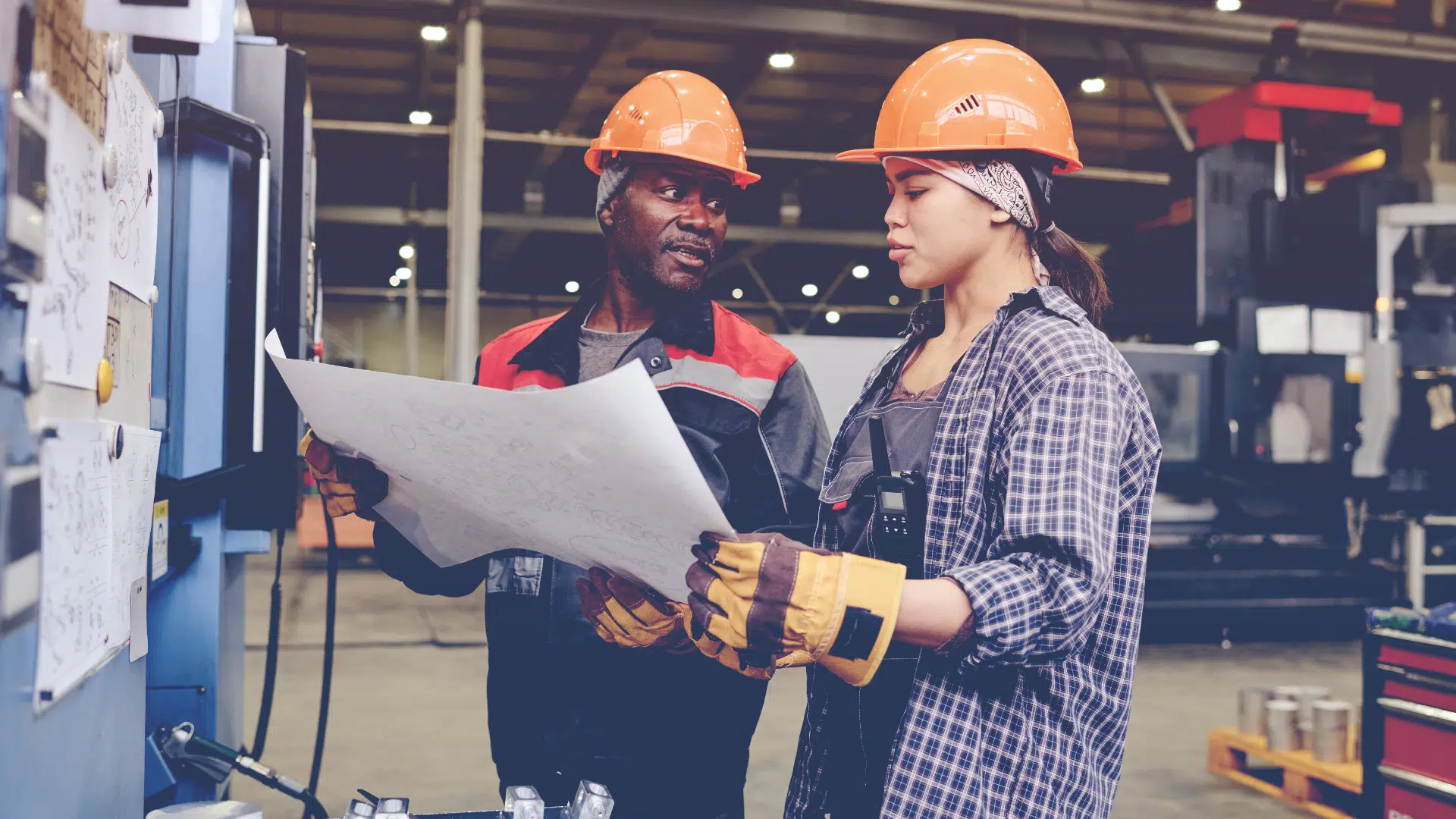two manufacturing workers in hard hats reading a blueprint and looking at machinery